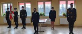 Komendant Powiatowy St. kpt. Tadeusz Kmieć podczas przemówienia z okazji ślubowania strażaka. W tle poczet sztandarowy, funkcjonariusz z sekcji kadr oraz dowódca uroczystości.