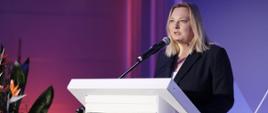 Wiceminister Ewelina Owczarska stoi przy mównicy i przemawia do mikrofonu