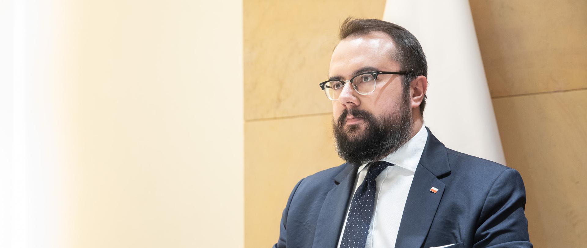 Mężczyzna z brodą, okularach i garniturze siedzi przy stole.