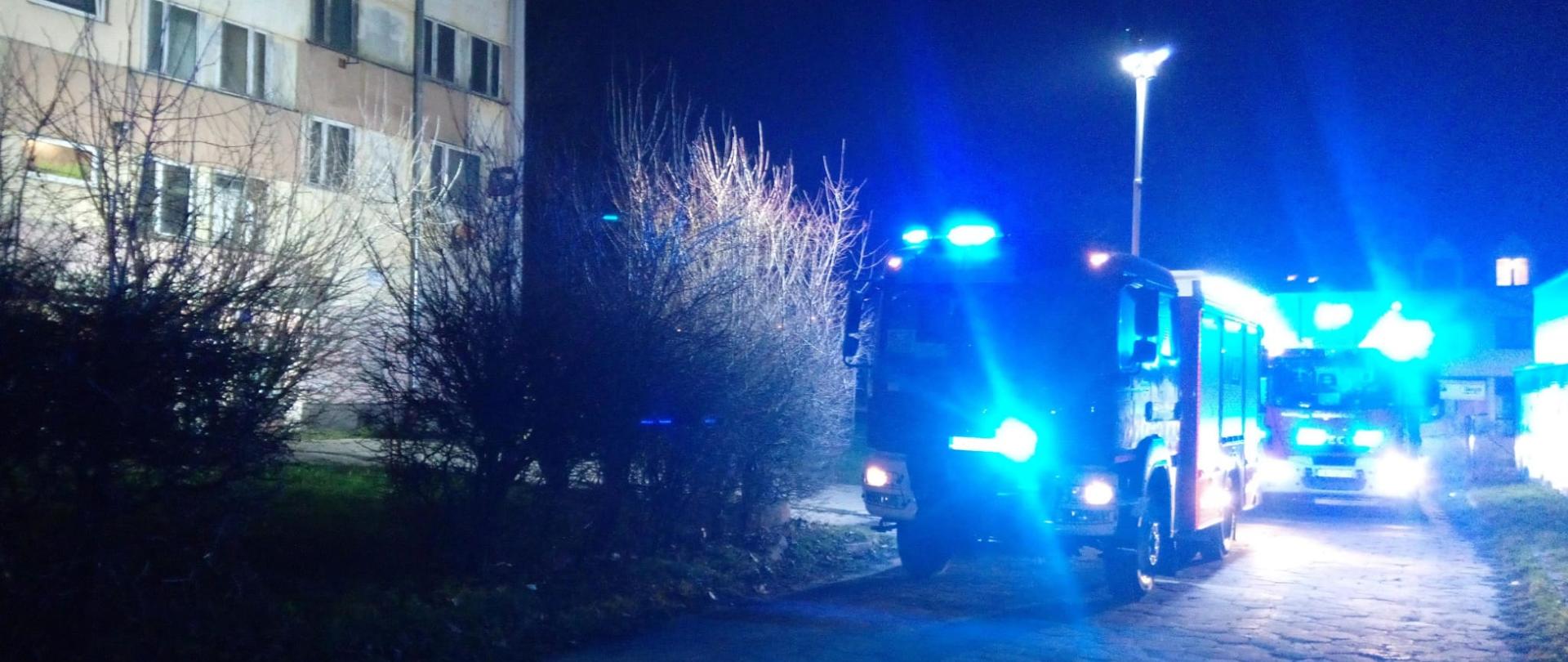 Zdjęcie przedstawia dwa samochody pożarnicze stojące przy budynku socjalnym, w którym doszło do pożaru. Budynek jest widoczny po lewej stronie.