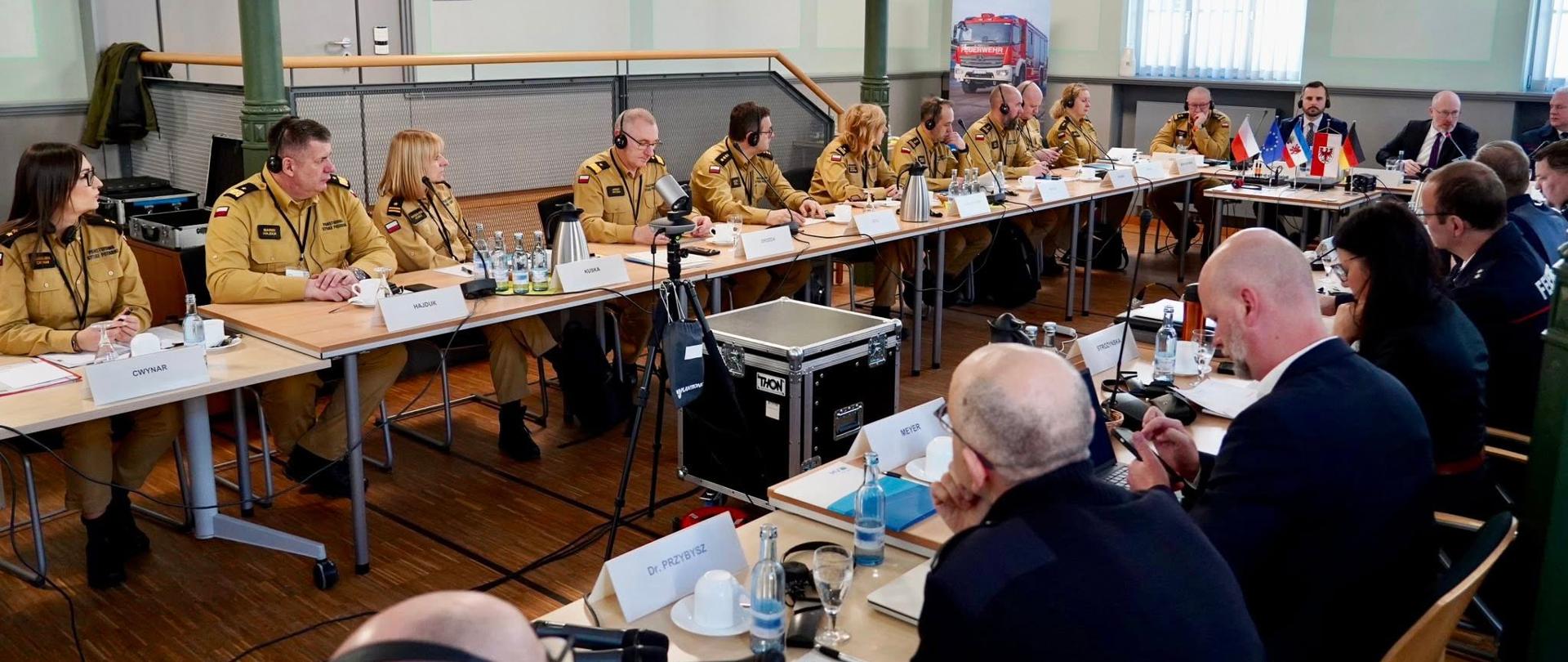 W przestronnej sali konferencyjnej odbywa się formalne spotkanie kilkunastu osób siedzących przy ustawionych w podkowę stołach. Większość uczestników to osoby w średnim wieku, ubrane w jasnobrązowe mundury służb ratowniczych lub straży pożarnej, z naszywkami i oznaczeniami na ramionach. Kilka osób po przeciwnej stronie stołu ma na sobie ciemne garnitury.
Na stołach znajdują się mikrofony, słuchawki do tłumaczeń symultanicznych, butelki wody, filiżanki, dokumenty oraz tabliczki z nazwiskami uczestników. Część osób korzysta ze słuchawek, inni robią notatki lub słuchają wypowiedzi. W centralnej części stoi statyw z kamerą lub sprzętem nagrywającym.
Na końcu sali widoczny jest roll-up z wizerunkiem czerwonego wozu strażackiego. Obok ustawione są małe flagi kilku krajów, co sugeruje międzynarodowy charakter spotkania. Pomieszczenie jest jasne, z wysokimi oknami i pionowymi żaluzjami oraz wiszącymi lampami sufitowymi. Atmosfera jest oficjalna i skupiona, a uczestnicy wydają się zaangażowani w dyskusję.