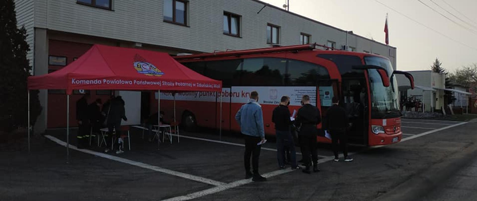 Na zdjęciu widoczny jest czerwony autokar Mobilny Punkt Poboru Krwi, Z lewej strony namiot z logiem PSP oraz napisem Komenda Powiatowa Państwowej Straży Pożarnej w Zduńskiej Woli pod którym rozstawione są stoliki i krzesełka. Przed wejściem do autokaru ustawionych jest kilka osób w kolejce. 