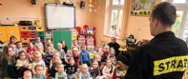 Strażak przedstawia przedszkolakom ubranie strażackie. Zdjęcie wykonane w budynku przedszkola.