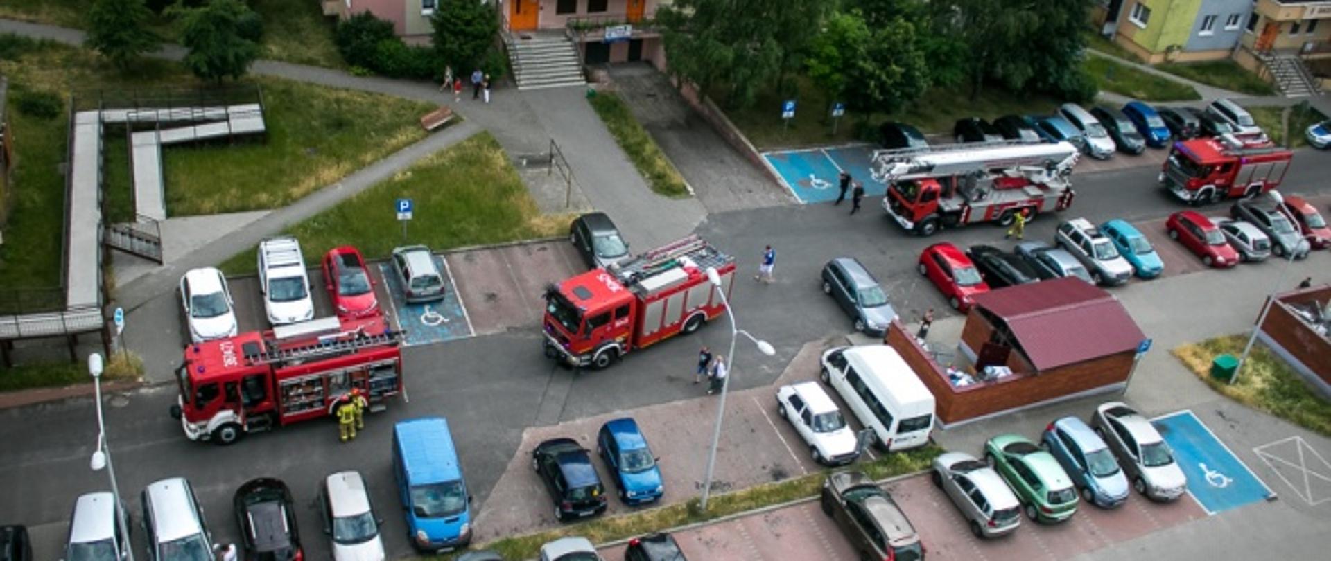 Zdjęcie przedstawia samochody strażackie na parkingu przed budynkiem mieszkalnym wielorodzinnym. Ponadto widać zaparkowane samochodu osobowe.