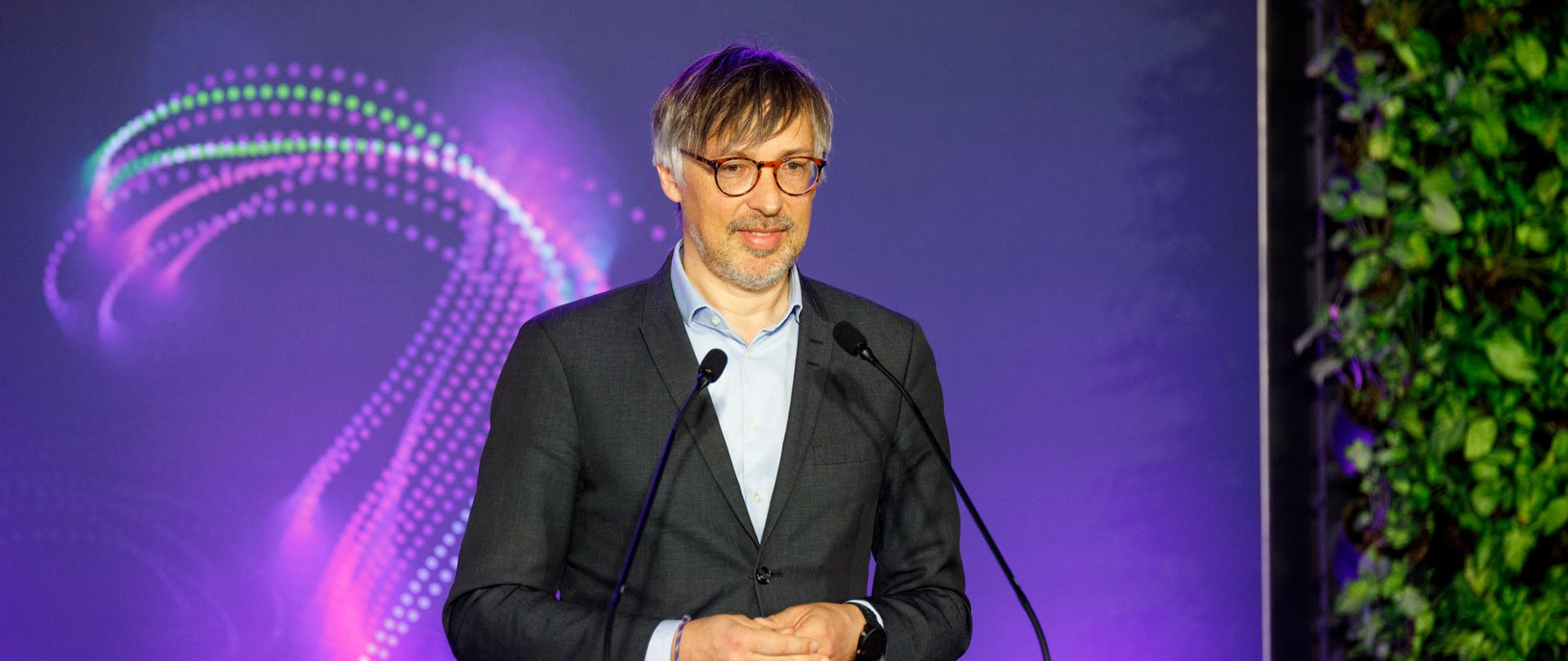 W centralnej części zdjęcia znajduje się wiceminister Krzysztof Bolesta. Stoi za mównicą. Za nim widoczny jest ekran, wyświetla się na nim fioletowe tło. Po prawej stronie zdjęcia widoczna jest roślinność, element scenografii. 