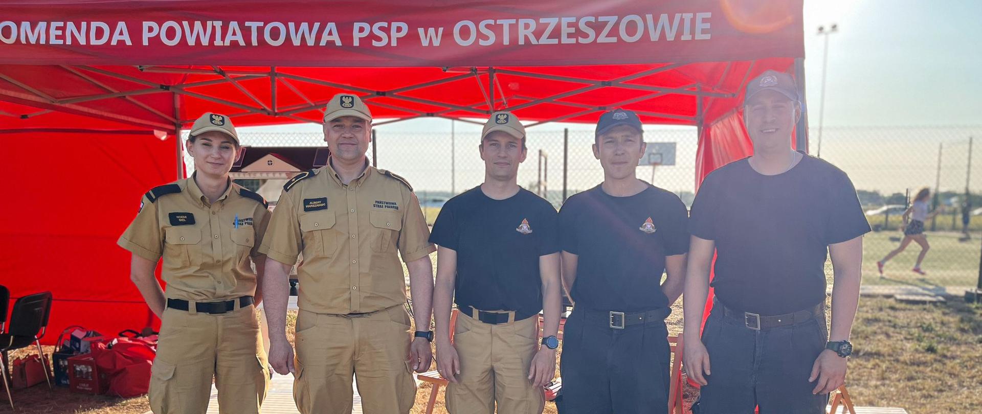 Strażacy biorący udział w akcji prewencyjnej. W tle ekspozycja pożarnicza.
