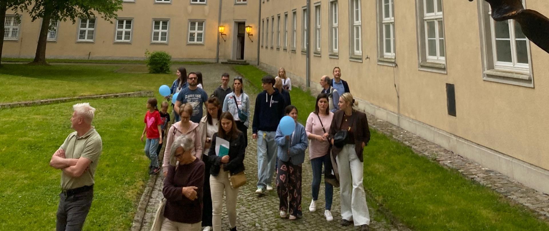 Dziedziniec otoczony jasnymi budynkami o klasycznej architekturze podczas pochmurnego dnia. Na pierwszym planie widoczna jest duża, stylizowana latarnia emitująca ciepłe żółte światło. W tle grupa osób spaceruje brukowaną ścieżką między trawnikiem a budynkiem; część uczestników trzyma niebieskie balony. Po lewej stronie rośnie duże zielone drzewo, a cała scena ma spokojny klimat.