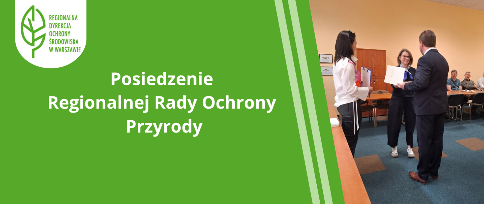 Mężczyzna w garniturze wręcza dokument kobiecie, kobieta w białej bluzce stoi z boku, w tle osoby siedzące za stołem
