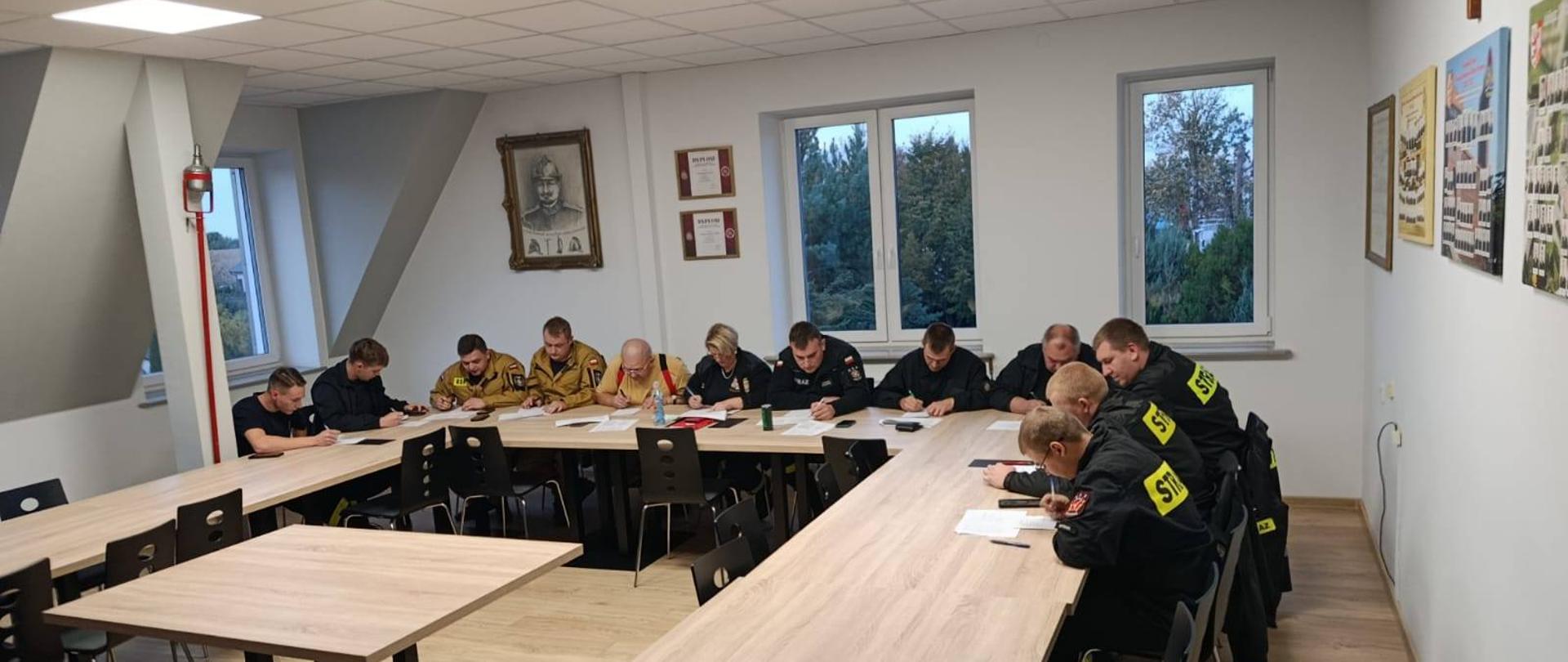Na zdjęciu widać grupę osób siedzących przy długich stołach, piszących egzamin. Wszyscy mają na sobie mundury strażackie. 