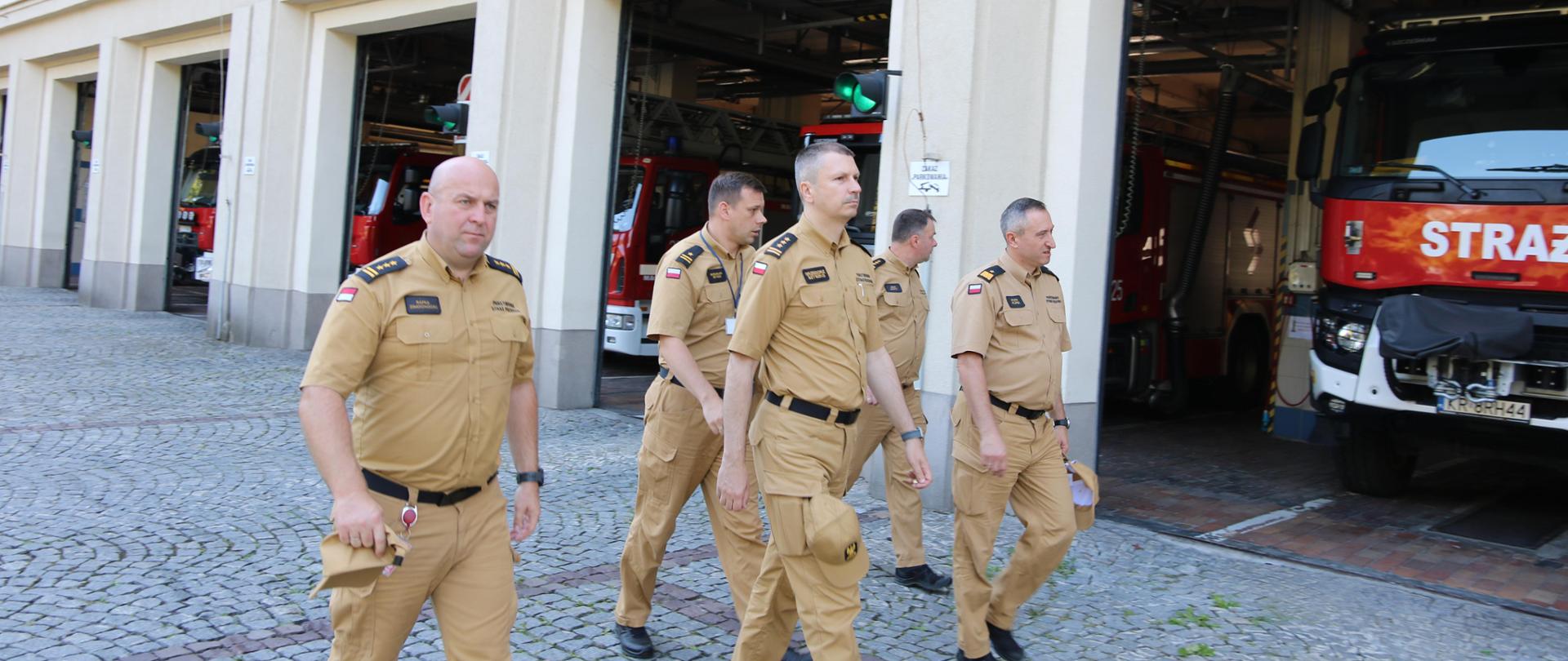 Plac wewnętrzny Szkoły Aspirantów PSP w Krakowie. Na nim 5 mężczyzn strażaków w umundurowaniu służbowym koloru piaskowego. W tle otwarte bramy garaży wewnątrz których zaparkowane są czerwonego koloru samochody ratowniczo-gaśnicze. 