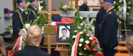 Trumna nakryta flagą biało - czerwoną, po obu stronach stoją strażacy , przed trumną ustawiona fotografia zmarłego strażaka