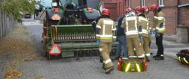 Wypadek podczas prac polowych. Teren jednego z zakładów pracy na terenie Rawicza. Sześciu strażaków na tle ciągnika rolniczego z podczepionym siewnikiem i budynków z czerwonej cegły, udziela kwalifikowanej pierwszej pomocy kobiecie z urazem ręki. Przed strażakami stoją dwie torby ze sprzętem ratownictwa medycznego.