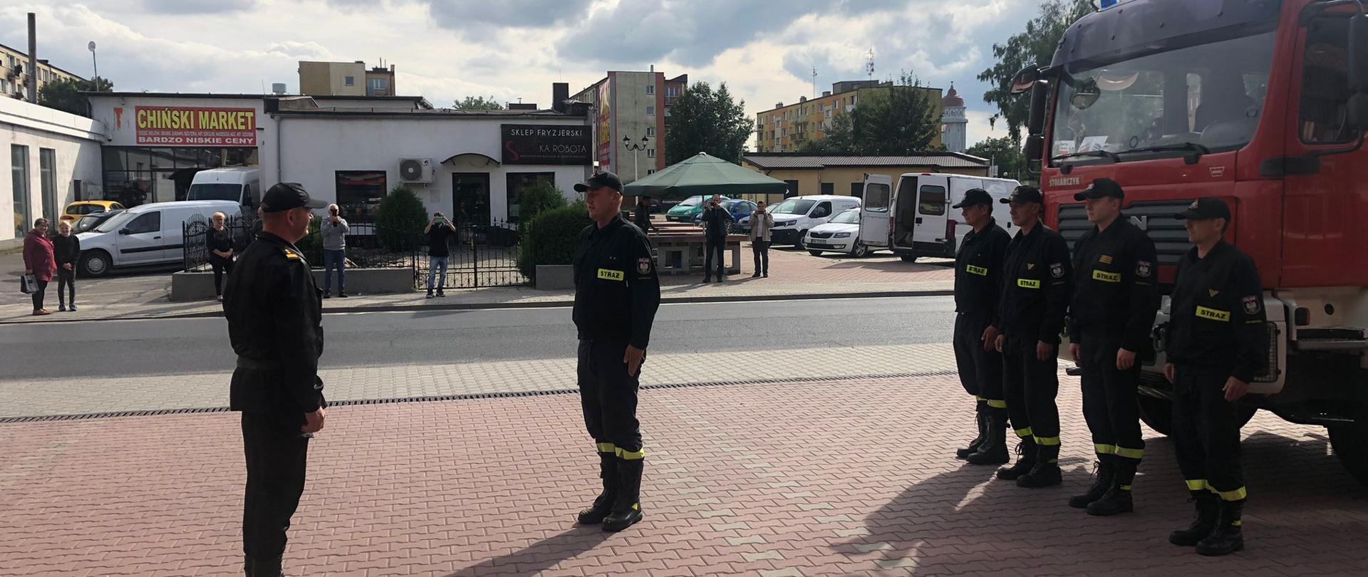 Zdjęcie przedstawia uroczyste powitanie strażaków na palcu zewnętrznym przed Komendą, którzy wrócili z misji gaszenia pożarów w Grecji. Na zdjęciu po prawej stronie widoczny zastęp, który wrócił z misji. Z lewej strony widzimy Komendanta PSP w Ostrzeszowie przyjmującego meldunek od dowódcy zastępy, który brał udział w misji gaszenia pożarów w Grecji. Zdjęcie wykonane w porze dziennej na placu wewnętrznym Komendy.