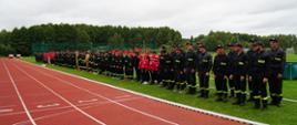 Długi szereg strażaków oraz członków młodzieżowych drużyn pożarniczych stojących w zbiórce na trawie przy bieżni lekkoatletycznej. Uczestnicy są ubrani w ciemne mundury strażackie i czerwone czapki.