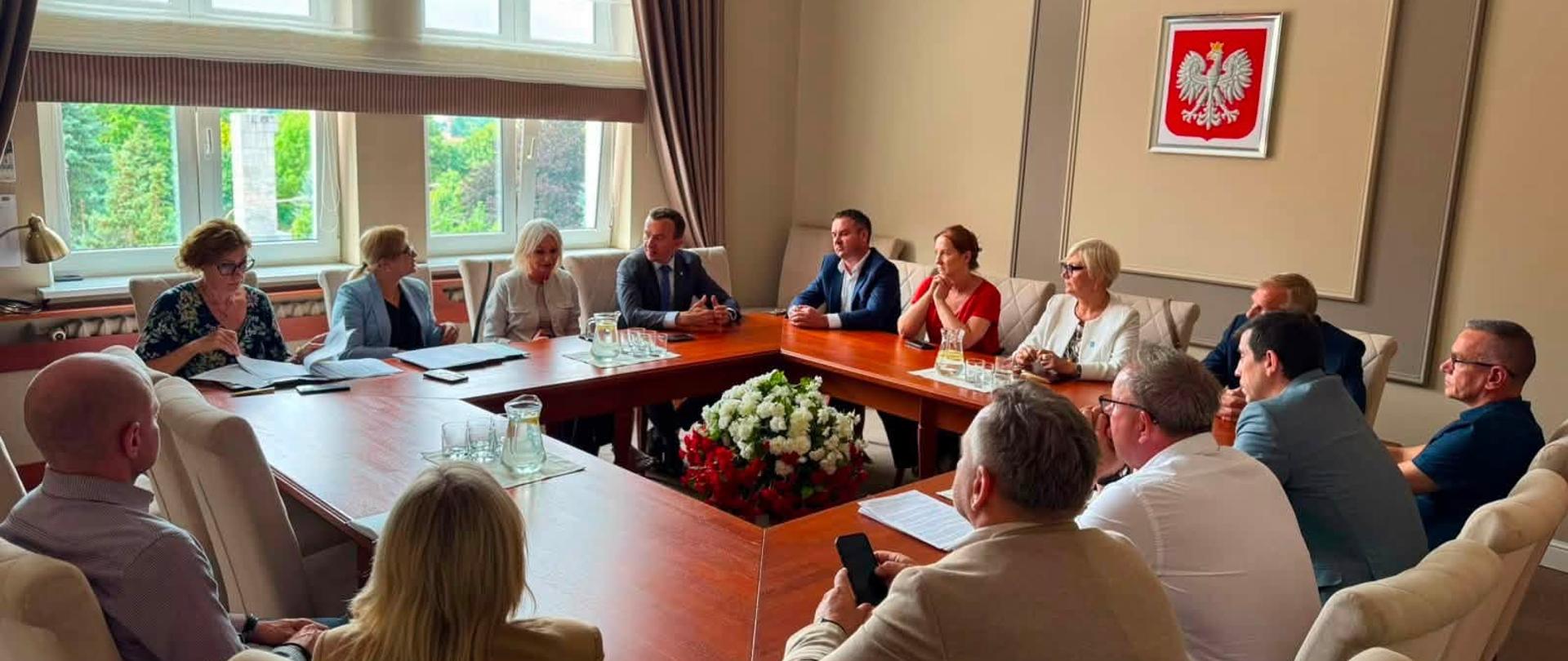 Grupa uczestników, w tym wicewojewoda, siedzi przy drewnianym stole konferencyjnym w jasnej sali. Na ścianie za nimi znajduje się godło Rzeczypospolitej Polskiej.