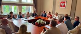 Grupa uczestników, w tym wicewojewoda, siedzi przy drewnianym stole konferencyjnym w jasnej sali. Na ścianie za nimi znajduje się godło Rzeczypospolitej Polskiej.