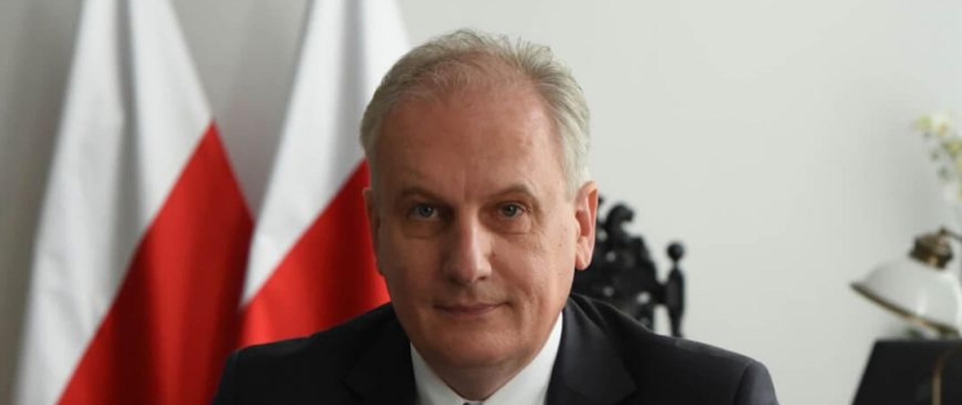 Na zdjęciu siedzący Wojewoda Pomorski w garniturze podpisuje umowe na przekazanie środków. Przed nim na stole biało-czerwony bukietr kwiatów. W tle flagi p[olski oraz godło