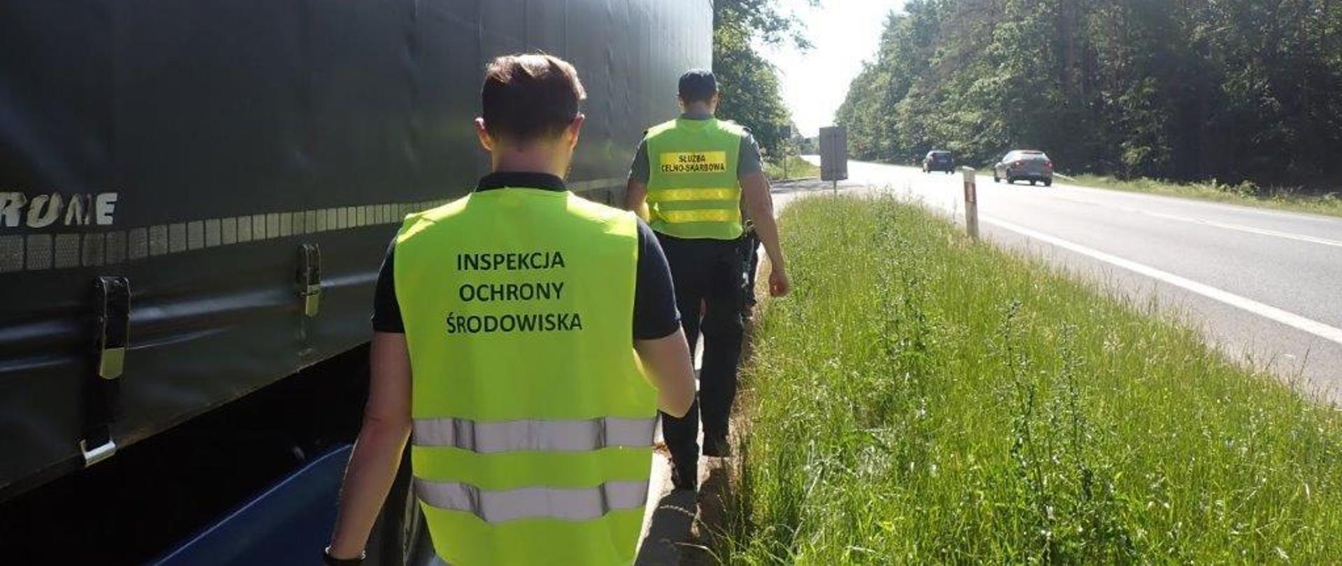 Pracownicy Inspekcji Ochrony Środowiska i Służby Celno-Skarbowej idą wzdłuż naczepy kontrolowanego pojazdu stojącego na parkingu przy drodze.