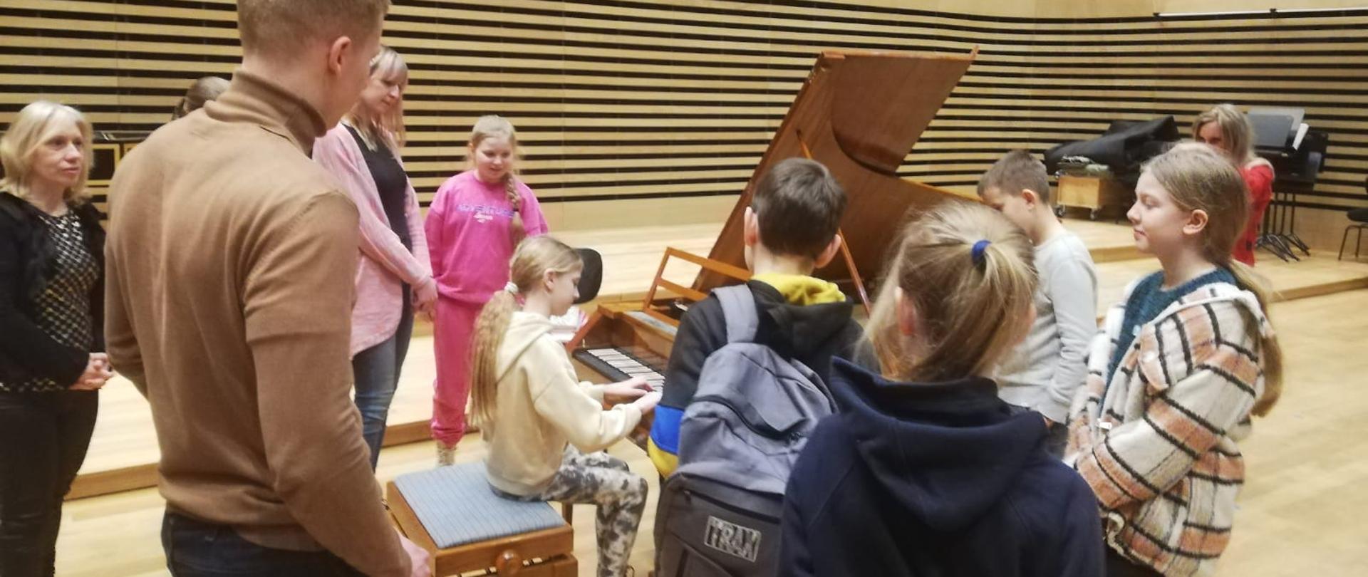 Dziewczynka gra na brązowym fortepianie z czarno-białą klawiaturą (kolory na odwrót jak we współczesnych fortepianach), dookoła ze wszystkich stron fortepianu stoi sześcioro dzieci i trójka dorosłych.