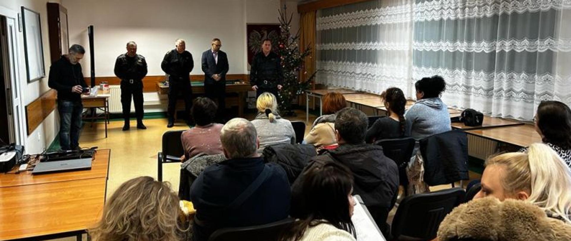 Na zdjęciu widać strażaków stojących na środku sali przed zebrynami gośćmi. Goście słuchają przemówienia Komendanta. W tle widać choinkę świąteczną.