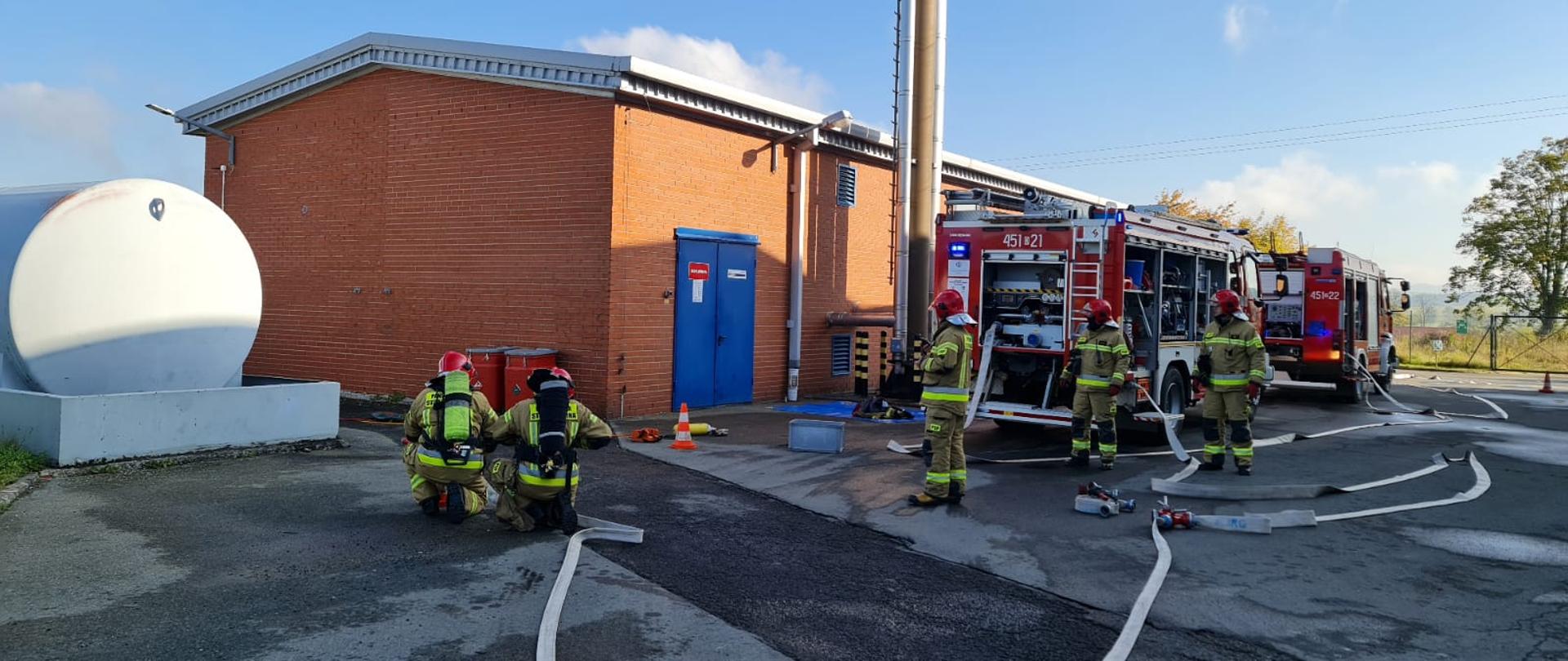Po prawej stronie widoczny walcowatego kształtu zbiornik na gaz obok niego dwóch ratowników przy odcinku ustawia kurtynę wodną po prawej stronie dwa samochody ratowniczo gańsnicze obok nich na ziemi znajdują się węże strażackie oraz rozdzielacz do którego podłączone są odcinki. za pojazdami znajduje się budynek zakładu produkcyjnego 