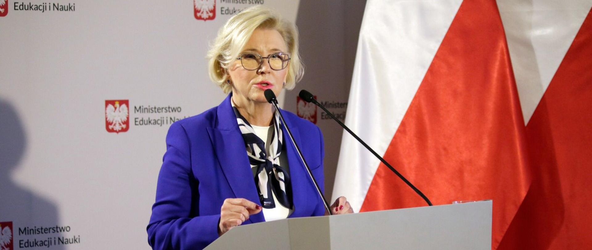 Wiceminister Marzena Machałek stoi przed mównicą podczas konferencji, za nią stoi baner z napisem Ministerstwo Edukacji i Nauki oraz flagi Polski.