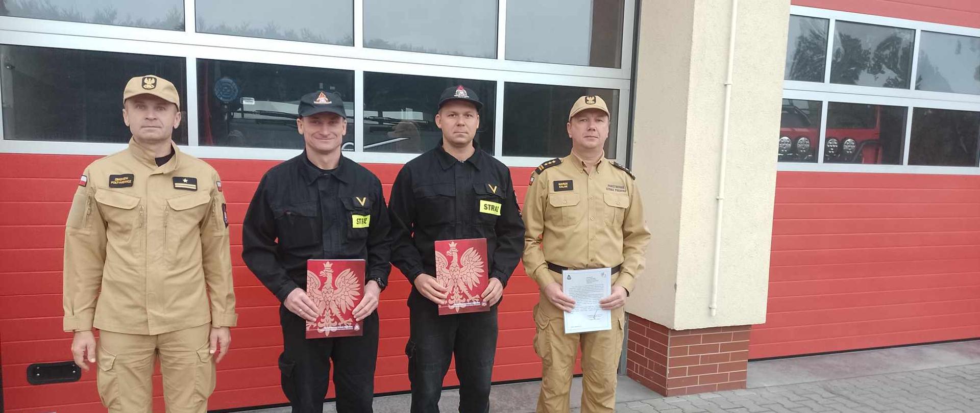 Zdjęcie przedstawia strażaków stojących na tle bramy garażowej koloru czerwonego. Dwóch strażaków stoi w umundurowaniu koloru żółtego (skrajni) oraz dwóch strażaków w umundurowaniu koloru czarnego (środkowi).