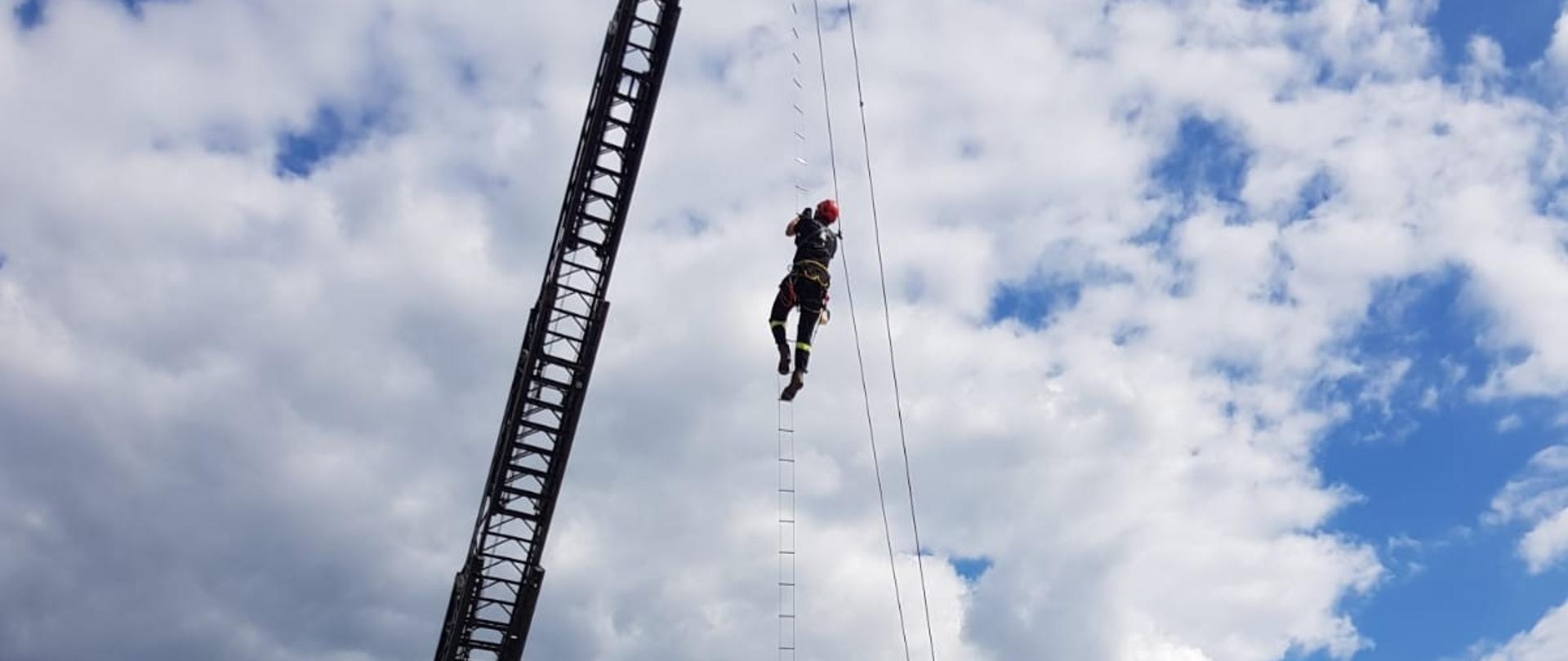 Strażak wspina się na drabince przywieszonej do kosza drabiny mechanicznej. W tle niebo z chmurami, w dole widać dachy samochodów strażackich i dach namiotu pneumatycznego. 