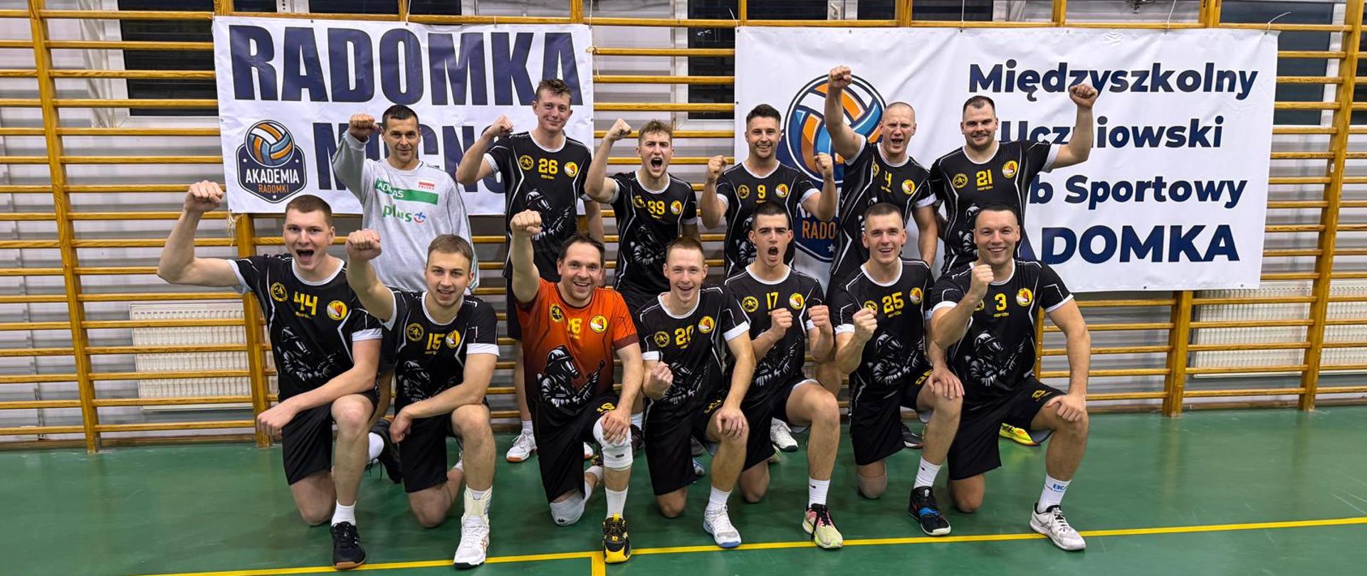 Zawodnicy drużyny siatkarskiej CS PSP wraz z trenerem na pamiątkowym zdjęciu, wszyscy uśmiechnięci z uniesioną w górę dłonią w geście triumfu, w tle wiszące na drabinkach banery klubu Radomka