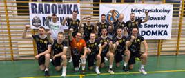 Zawodnicy drużyny siatkarskiej CS PSP wraz z trenerem na pamiątkowym zdjęciu, wszyscy uśmiechnięci z uniesioną w górę dłonią w geście triumfu, w tle wiszące na drabinkach banery klubu Radomka