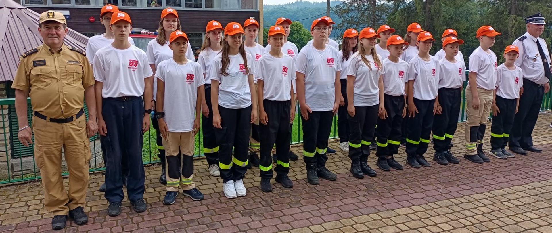 Zdjęcie zrobione na zewnątrz, podczas uroczystej zbiórki. W szeregu stoją uczestnicy obozu szkoleniowo-wypoczynkowego młodzieżowych drużyn pożarniczych, razem z nimi stoi oficer Państwowej Straży Pożarnej w stopniu starszego brygadiera.