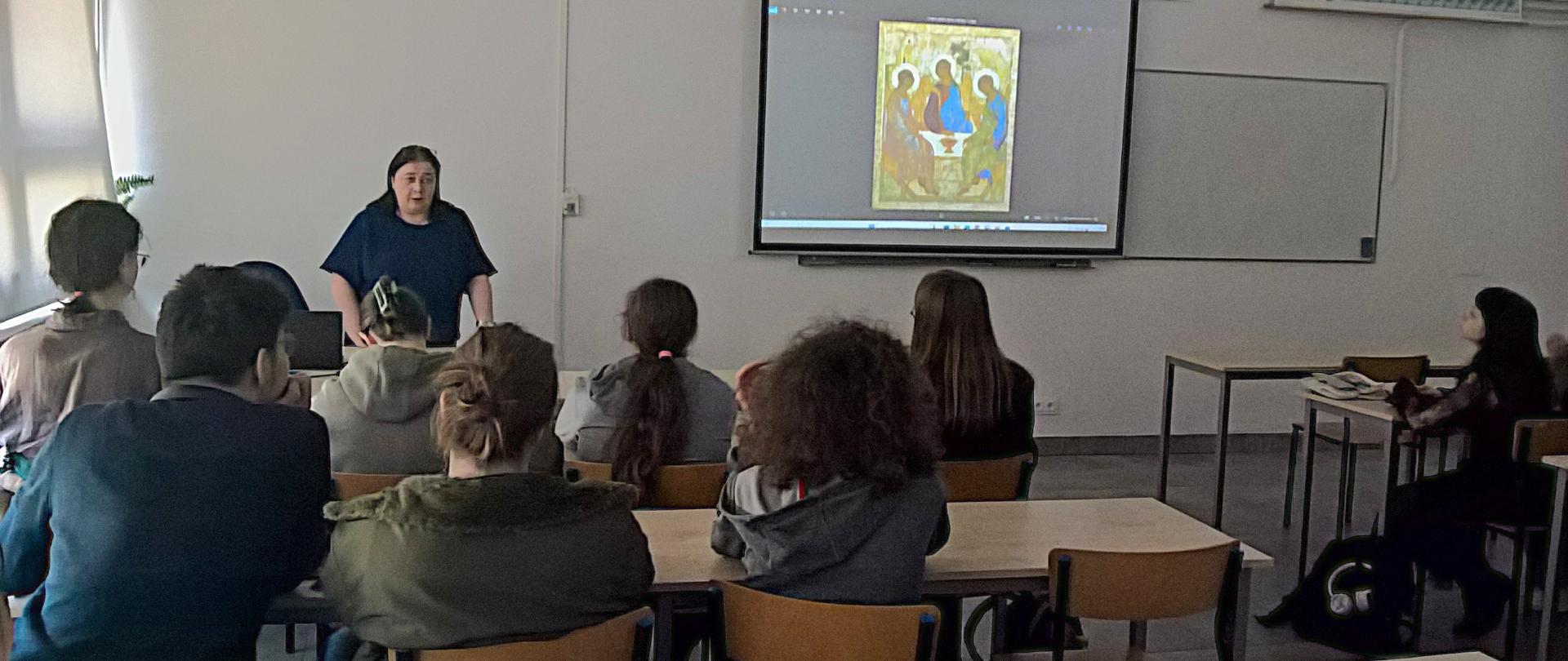 Anna Palusińska prowadzi wykład z uczniami w sali lekcyjnej PLSP w Lublinie. Uczniowie siedzą w ławce. Na tablicy wyświetlany jest slajd przedstawiający ikonę.