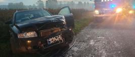 czarny samochód z rozbitym przodek stoi na poboczu drogi na drodze za nim samochód strażacki 