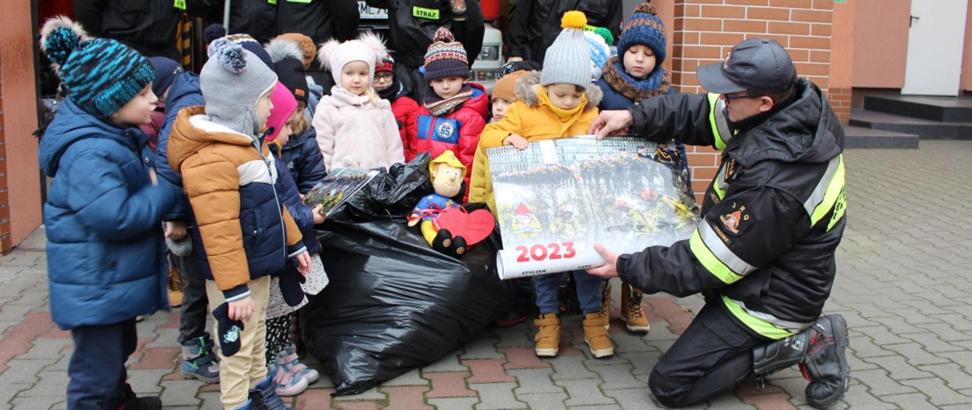 Zdjęcie przedstawia strażaków i dzieci w trakcie odwiedzania strażnicy.
W tle budynki.
