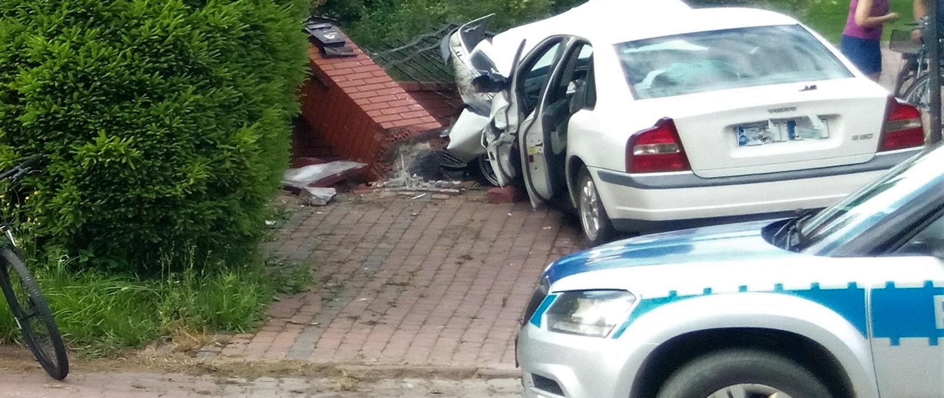 Na zdjęciu przedstawiono tył samochodu osobowego, który uderzył w ogrodzenie. Przed autem widać przewrócony murowany z klinkieru słupek ogrodzeniowy a bliżej patrzącego pokazano fragment policyjnego radiowozu.