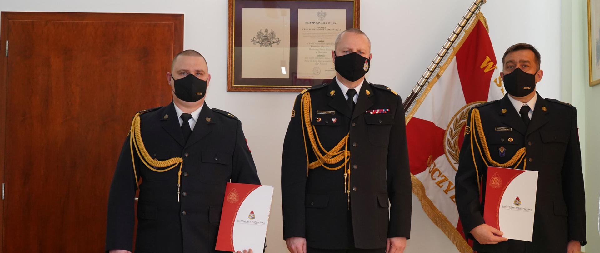 trzech strażakó stoi obok siebie, dwóch z nich trzyma teczkę, w tle sztandar