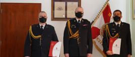 trzech strażakó stoi obok siebie, dwóch z nich trzyma teczkę, w tle sztandar