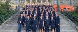 Konferencja kończąca program czesko-polski - wspólne zdjęcie wszystkich uczestników konferencji. Strażacy czescy i polscy w mundurach wyjściowych stoją na schodach obiektu, w którym odbywała się konferencja. Nad głowami uczestników powiewają flagi wielu państw.