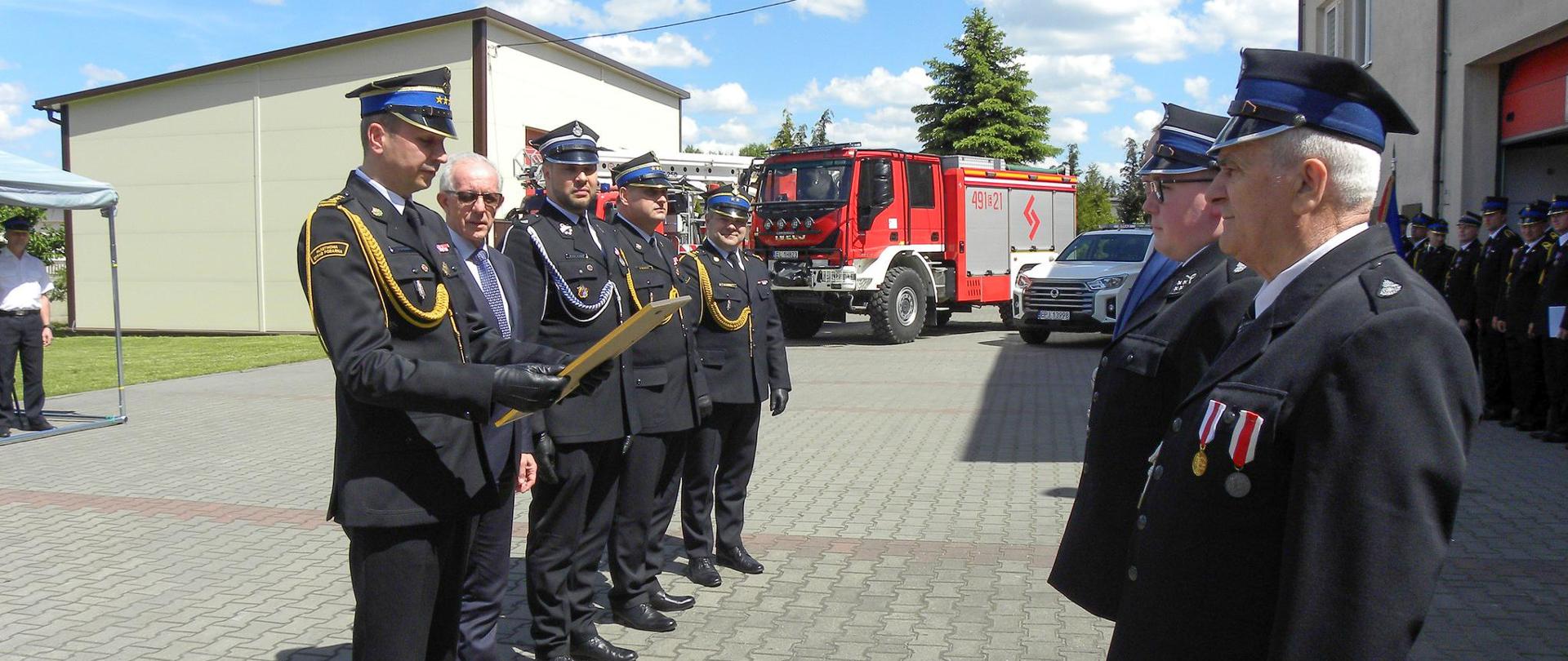 uroczystość na placu wewnętrznym komendy po lewej stoi czterech strażaków i mężczyzna w garniturze po prawej dwóch druhów w tle samochód pozarniczy i budynki komendy