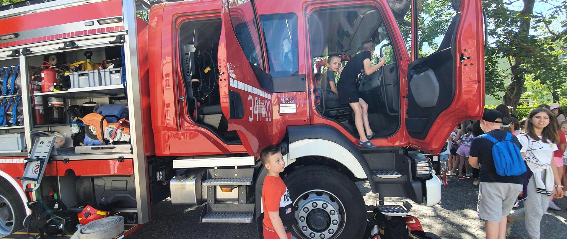 Na zdjęciu widać samochód ratowniczo-gaśniczy w kolorze czerwonym. Światła ostrzegawcze niebieskie. W pobliżu auta dzieci. Za autem teren zielony. Zdjęcie wykonane w ciągu dnia. Auto stoi na asfalcie.