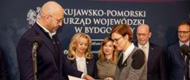 Zdjęcie z konferencji prasowej. Minister sportu i turystyki przekazuje decyzję o dofinansowaniu budowy lodowisk zastępczyni wójta Gminy Zławieś Wielka Joannie Wałeckiej. Za nimi stoją inni uczestnicy konferencji. W tle grantowa ścianka z napisem "Kujawsko-pomorski Urząd Wojewódzki w Bydgoszczy"