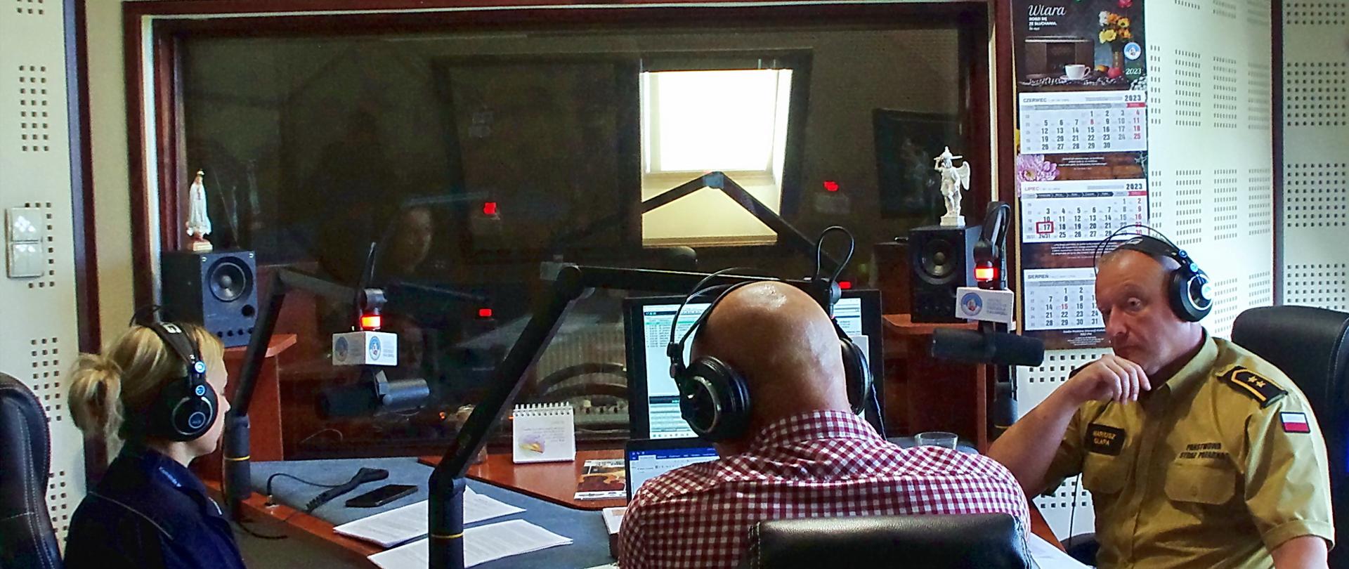W studiu radiowym siedzi strażak w żółtym mundurze państwowej straży pożarnej, policjantka w niebieskim mundurze policji oraz mężczyzna ubrany w kracistą koszulę, wszyscy mają słuchawki na uszach, strażak i policjantka słuchają swojego rozmówcę