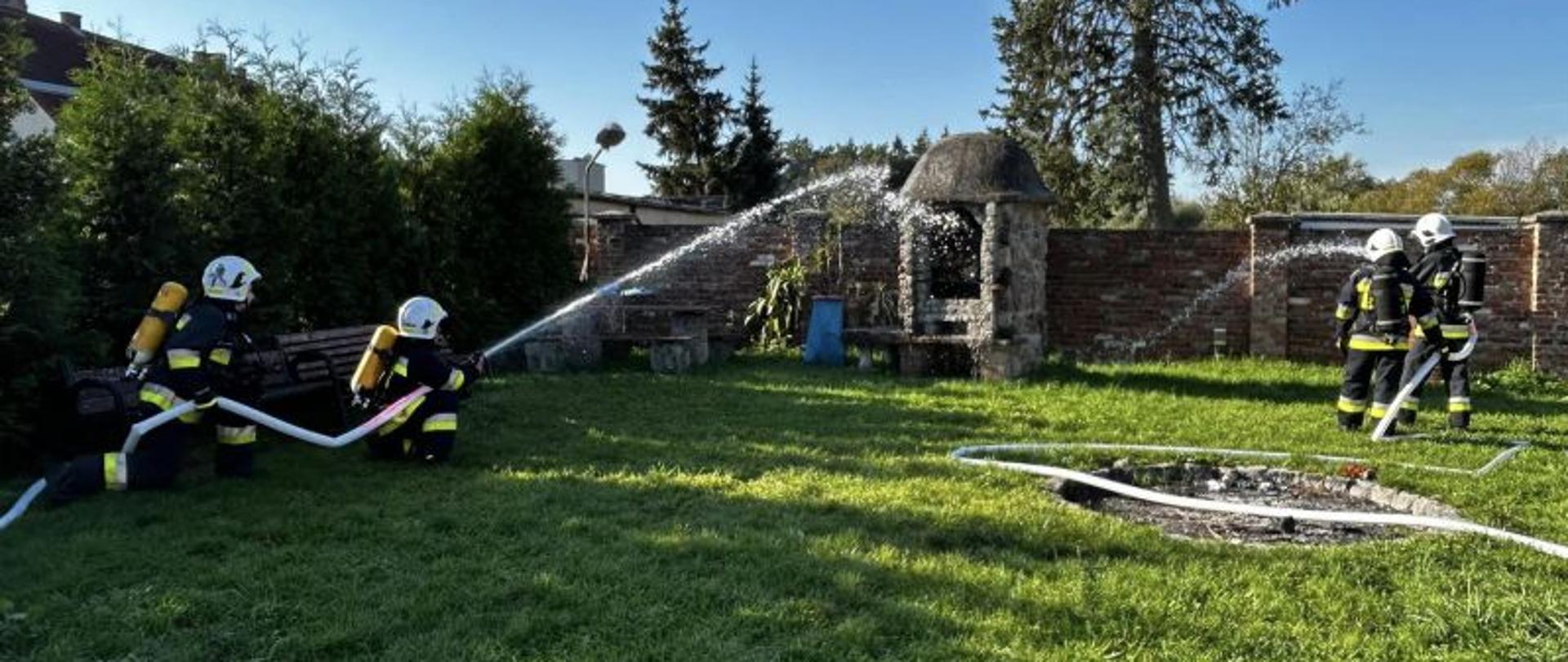 Widok na strażaków ćwiczących gaszenie. Po lewej stronie dwaj strażacy z aparatami ochrony dróg oddechowych podają wodę na duży murowany grill symulujący źródło pożaru. Po prawej również dwóch strażaków podających prąd wody na ten sam obiekt. Strażacy ćwiczą gaszenie.