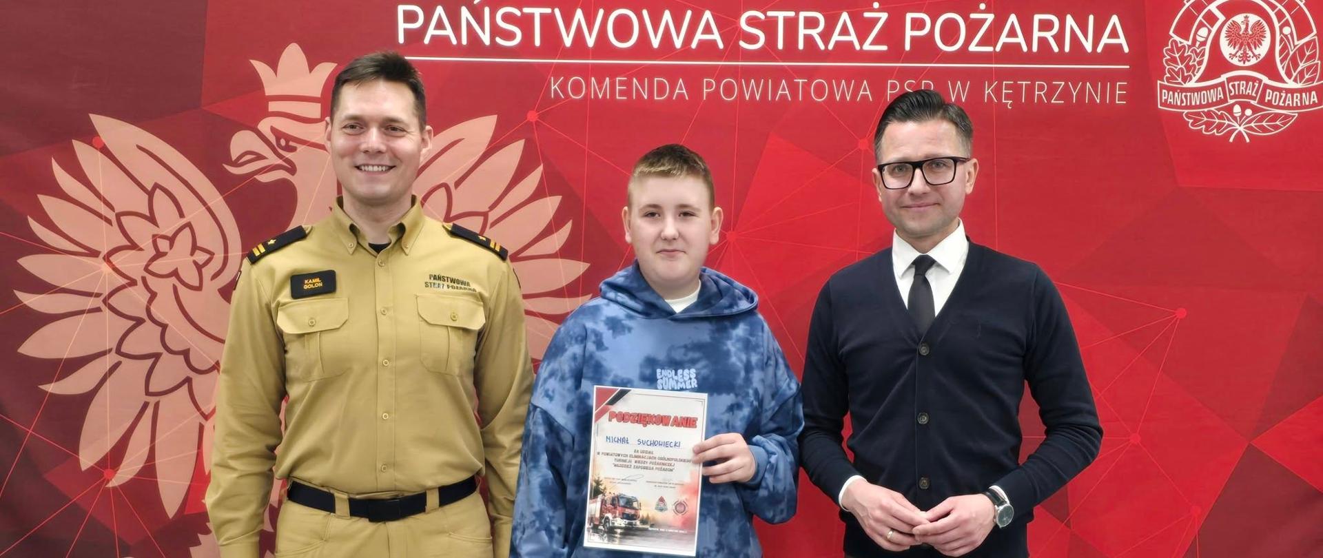 Na zdjęciu widzimy Starostę Powiatu Kętrzyńskiego oraz Komendanta Powiatowego Państwowej Straży Pożarnej w Kętrzynie na tle czerwonego banneru. W środku z nagrodą i dyplomem stoi uczeń.