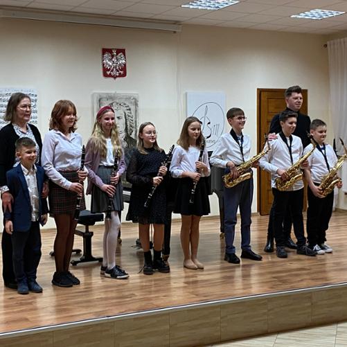 Zdjęcie przedstawia scenę, na scenie ośmioro uczniów z klarnetami i saksofonami. Skrajnie po lewej stoi akompaniator Iwona Czech. Skrajnie po prawej nauczyciel saksofonu i klarnetu Mateusz Kurkowski. W tle na ścianie portret J. S. Bacha oraz obraz z nutami i logiem szkoły. 