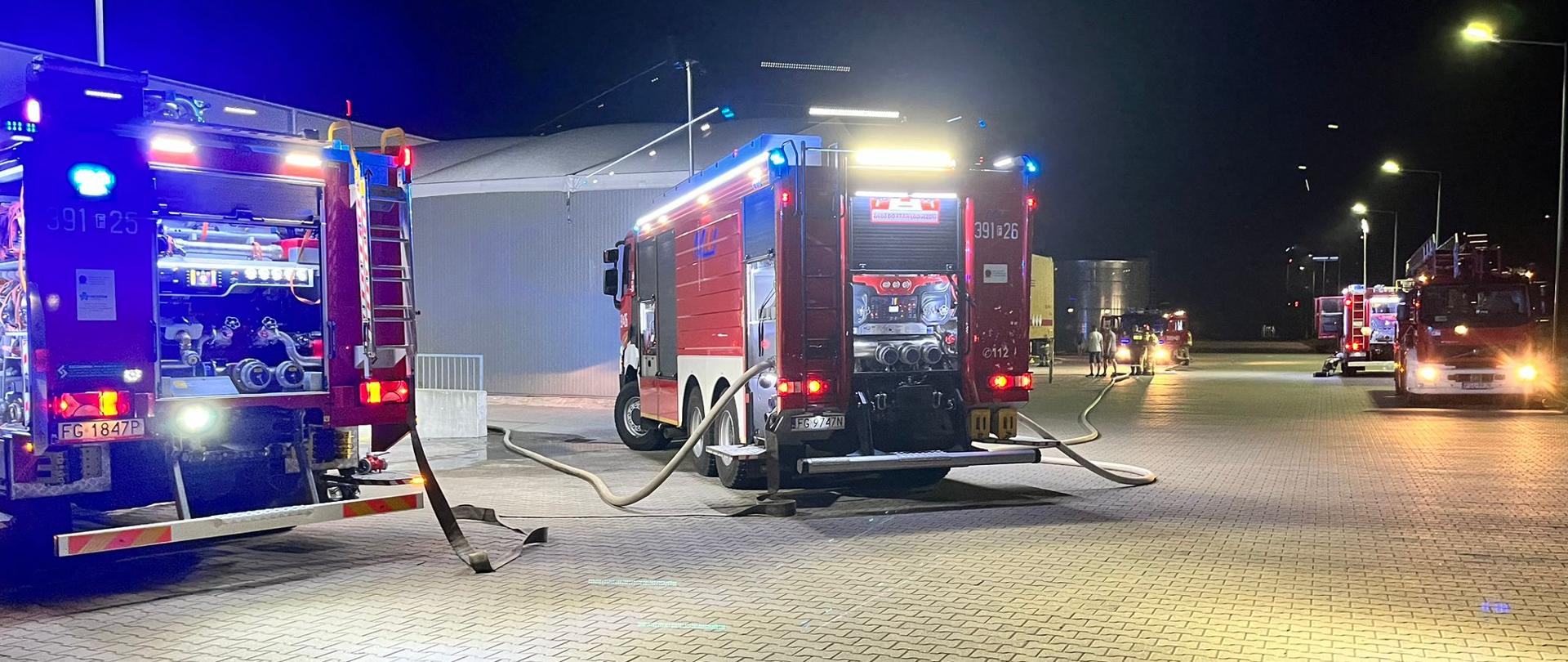 Samochody pożarnicze stojące na placu centrum logistycznego przed halą magazynową. Widoczne rozłożone i napełnione wodą węże strażackie.