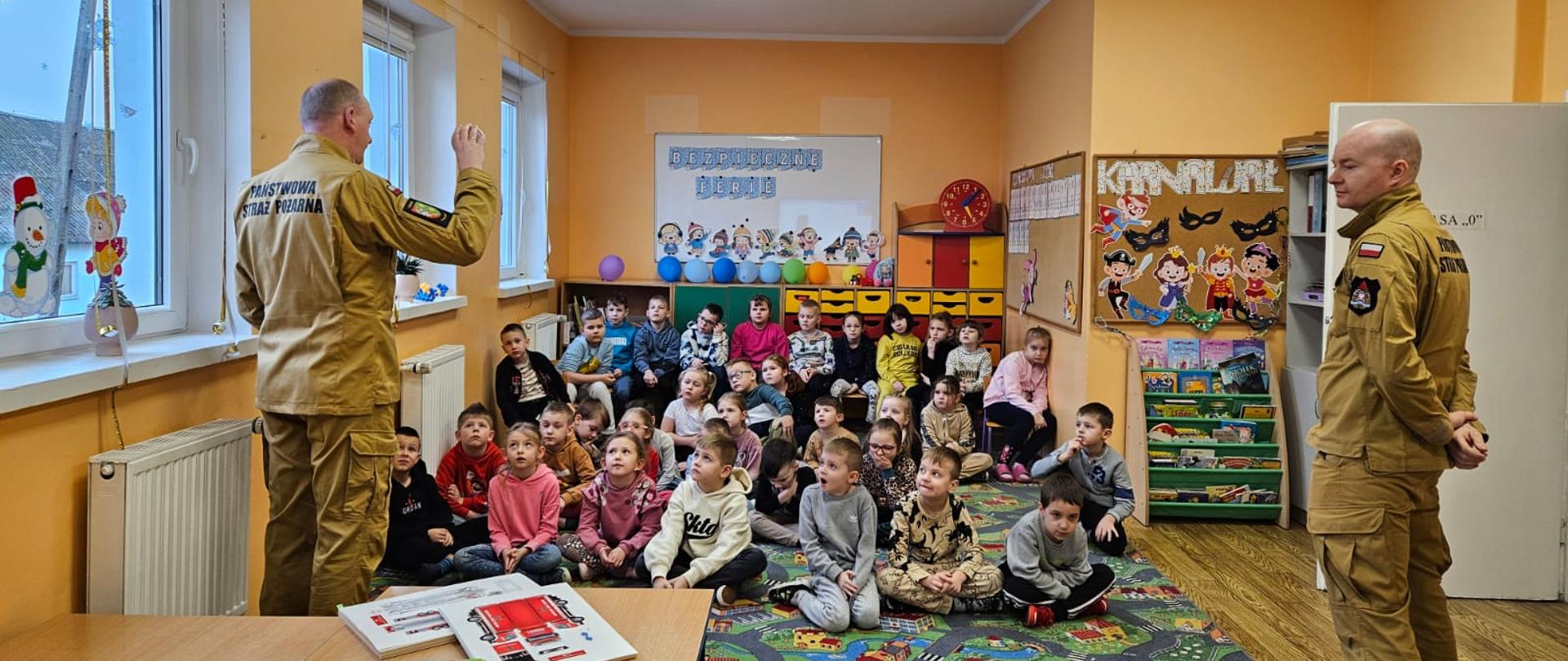 Dwóch umundurowanych strażaków stojących w sali przed grupą dzieci. Jeden ze strażaków prezentuje czujkę tlenku węgla.