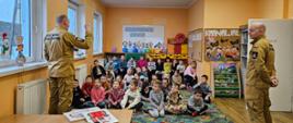 Dwóch umundurowanych strażaków stojących w sali przed grupą dzieci. Jeden ze strażaków prezentuje czujkę tlenku węgla.