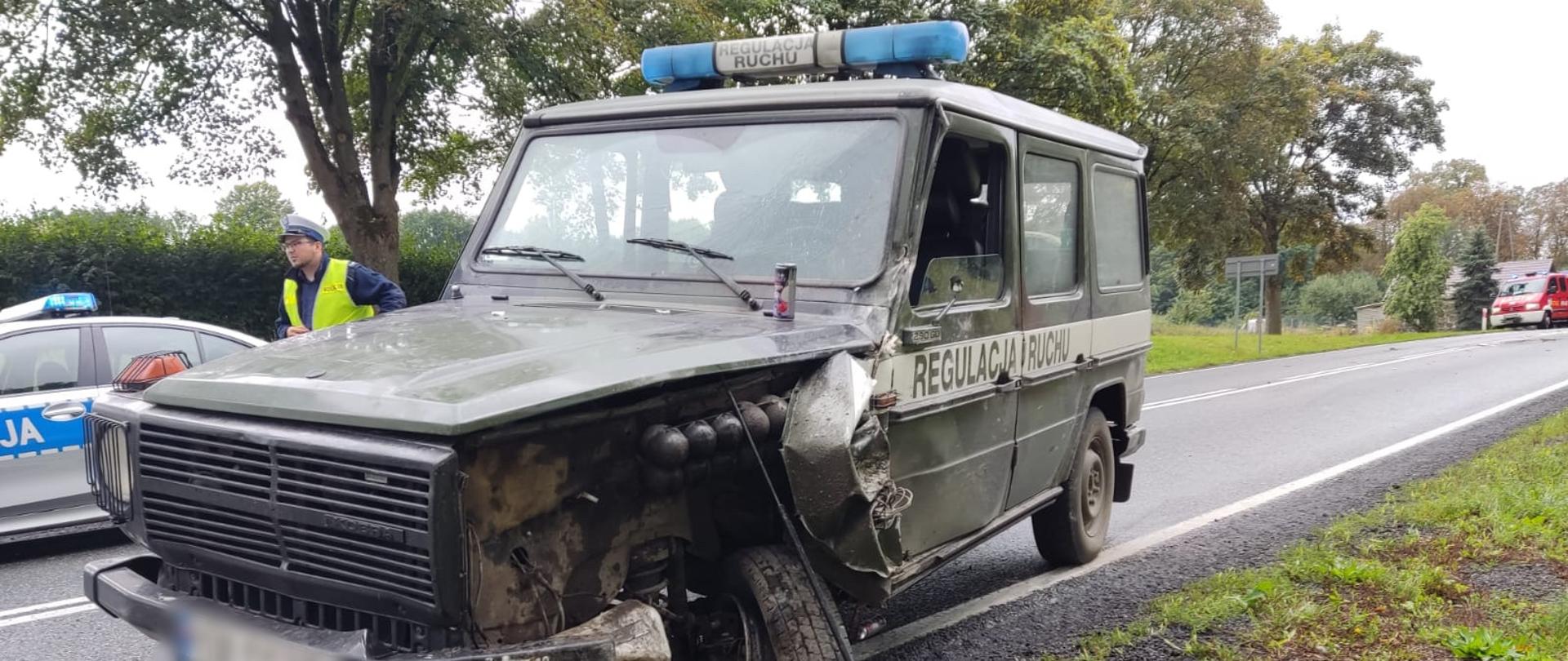 Na fotografii znajduje się wojskowy pojazd terenowy z niebieską belką świetlną na dachu. Pojazd jest pomalowany w barwach wojskowych. Pojazd ma rozbity lewy przód od strony kierowcy i uszkodzone koło. Pojazd znajduje się na pasie jezdni. Za pojazdem po lewej stronie znajduje się radiowóz, obok którego stoi policjant. Dookoła drogi rosną drzewa. W oddali po prawej stronie znajduje się wóz strażacki. Zdjęcie wykonano w porze dziennej przy pochmurnym niebie.