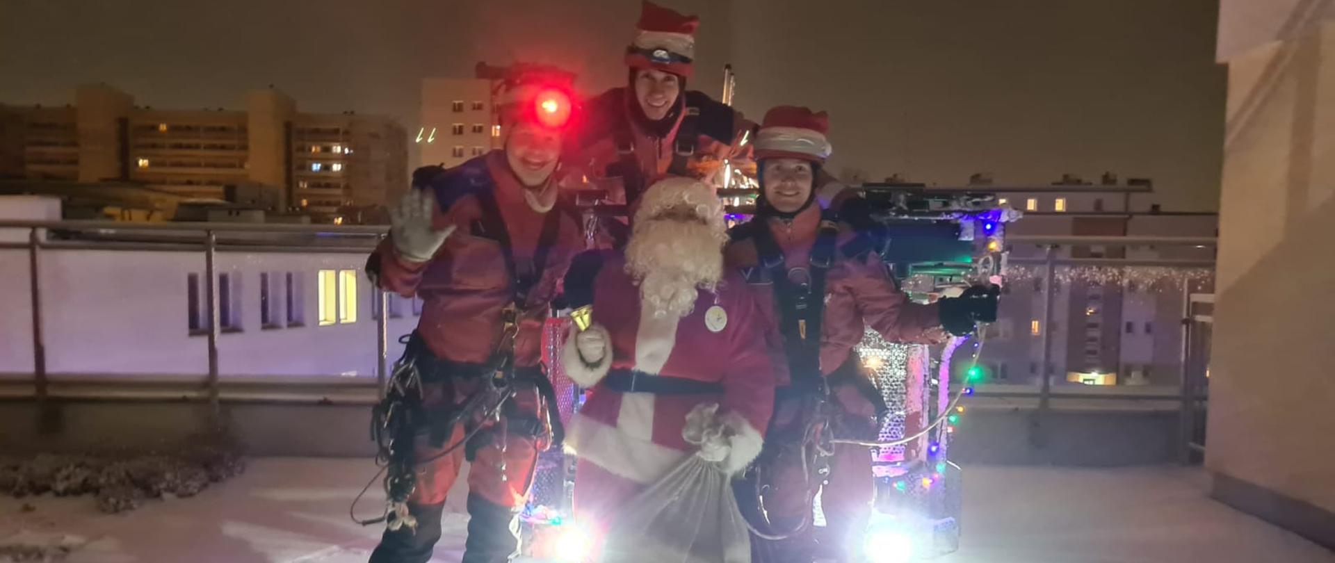 Trzej strażacy w czerwonych ubraniach grupy wysokościowej, w środku Święty Mikołaj, wszyscy stoją na balkonie budynku szpitala